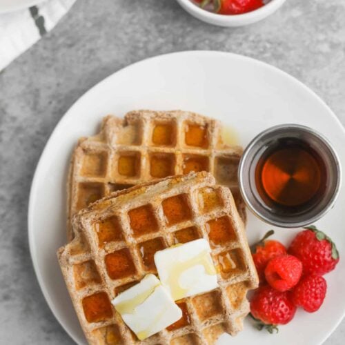 Whole Wheat Waffles 13 Two Whole Wheat Waffles With Butter And Syrup Rest On A White Plate, Accompanied By A Small Dish Of Syrup And A Few Strawberries. Nearby, Another Bowl Brims With Strawberries. The Backdrop Is A Textured Gray Surface, Creating A Cozy Breakfast Setting.