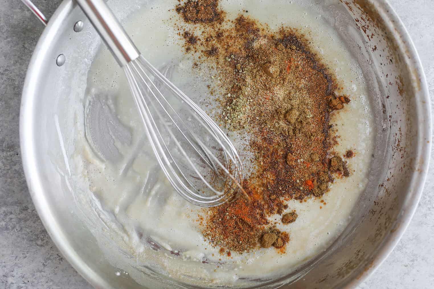 Bean and Cheese Enchiladas 3 A Metal Whisk In A Stainless Steel Pan Mixing Melted Butter With Spices And Seasonings, Creating A Roux For Bean And Cheese Enchiladas On A Gray Countertop.