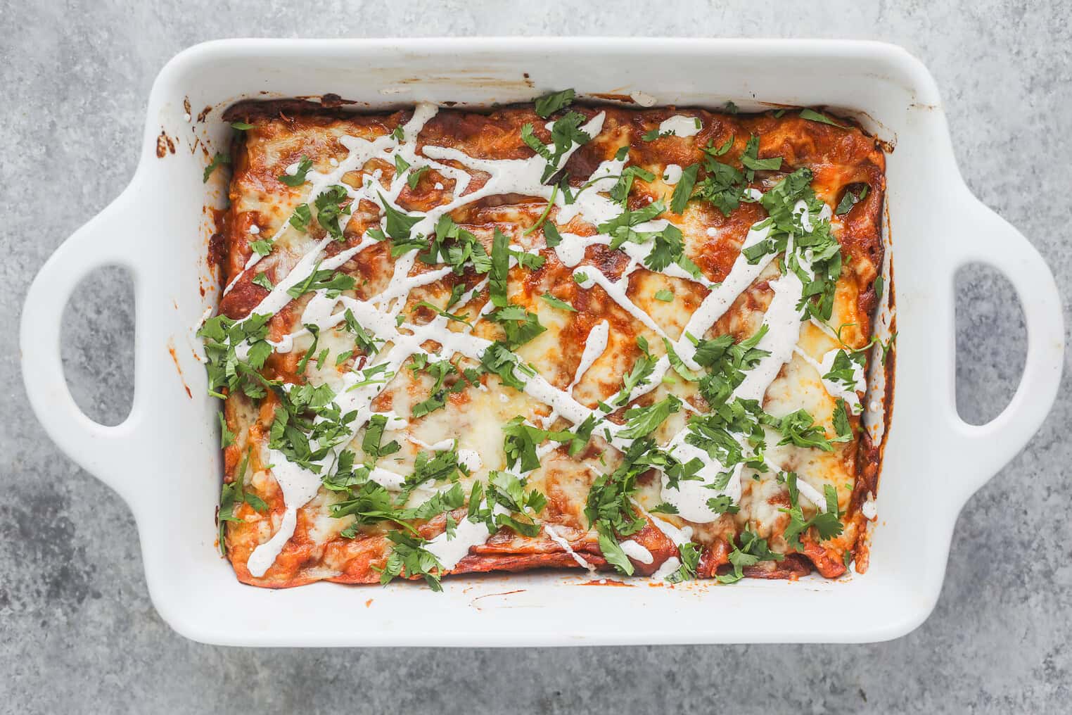 Bean and Cheese Enchiladas 11 A Baked Casserole Dish Filled With Bean And Cheese Enchiladas, Topped With Melted Cheese, Drizzled With White Sauce, And Garnished With Chopped Fresh Cilantro On A Gray Surface.