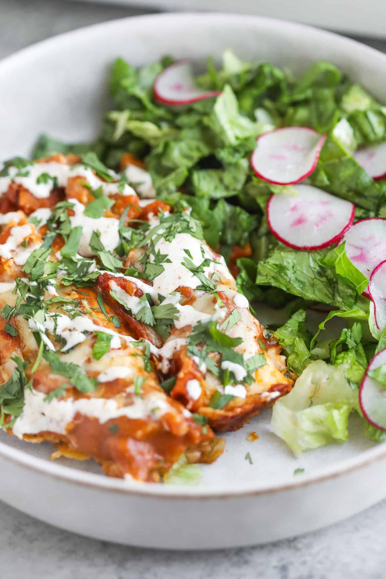 Bean and Cheese Enchiladas 10 Two Bean And Cheese Enchiladas Topped With Sauce, Crema, And Chopped Cilantro Are Served On A Plate Alongside A Fresh Green Salad With Sliced Radishes.