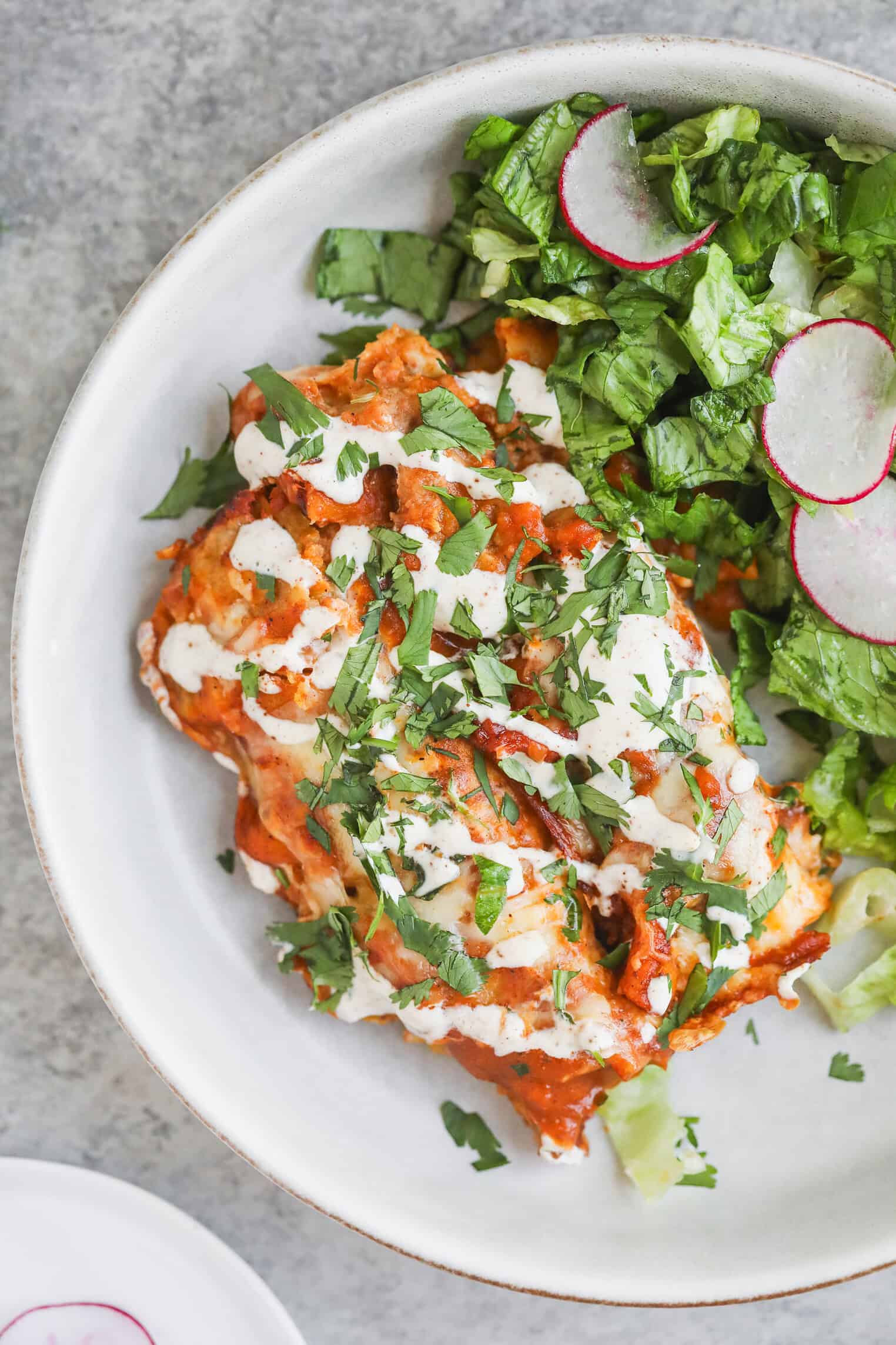A White Plate With Bean And Cheese Enchiladas Topped With Sauce, Crema, And Chopped Cilantro, Served With A Side Salad Of Lettuce And Sliced Radishes.