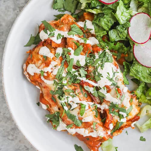 Bean and Cheese Enchiladas 14 A White Plate With Bean And Cheese Enchiladas Topped With Sauce, Crema, And Chopped Cilantro, Served With A Side Salad Of Lettuce And Sliced Radishes.