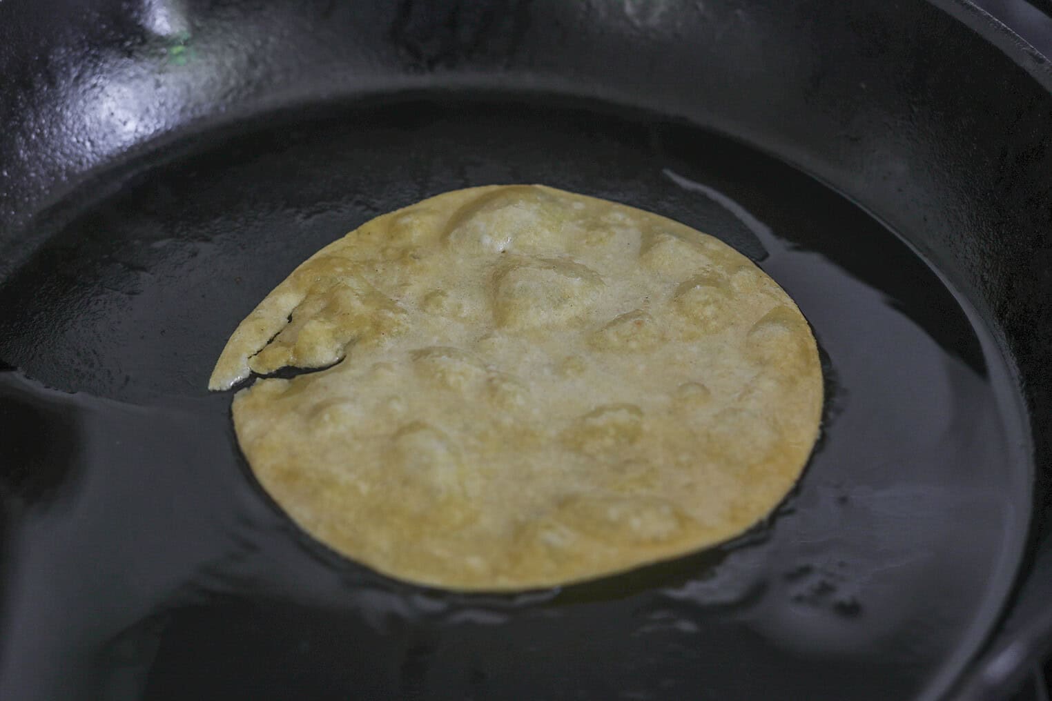 Bean and Cheese Enchiladas 5 A Corn Tortilla, Perfect For Bean And Cheese Enchiladas, Is Frying In Oil In A Black Skillet, With Bubbles Forming On Its Surface And Slightly Crisped Edges.