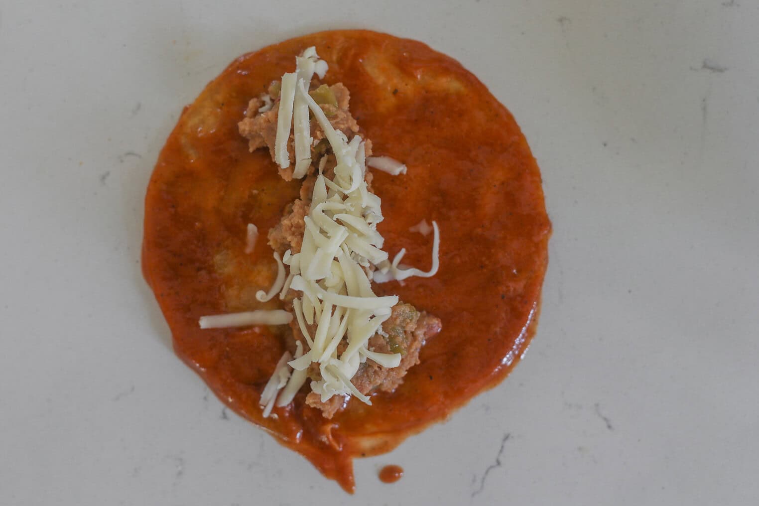 Bean and Cheese Enchiladas 6 A Corn Tortilla Topped With Red Sauce, Shredded Cheese, And Refried Beans Sits On A Light-Colored Countertop, Resembling Classic Bean And Cheese Enchiladas.