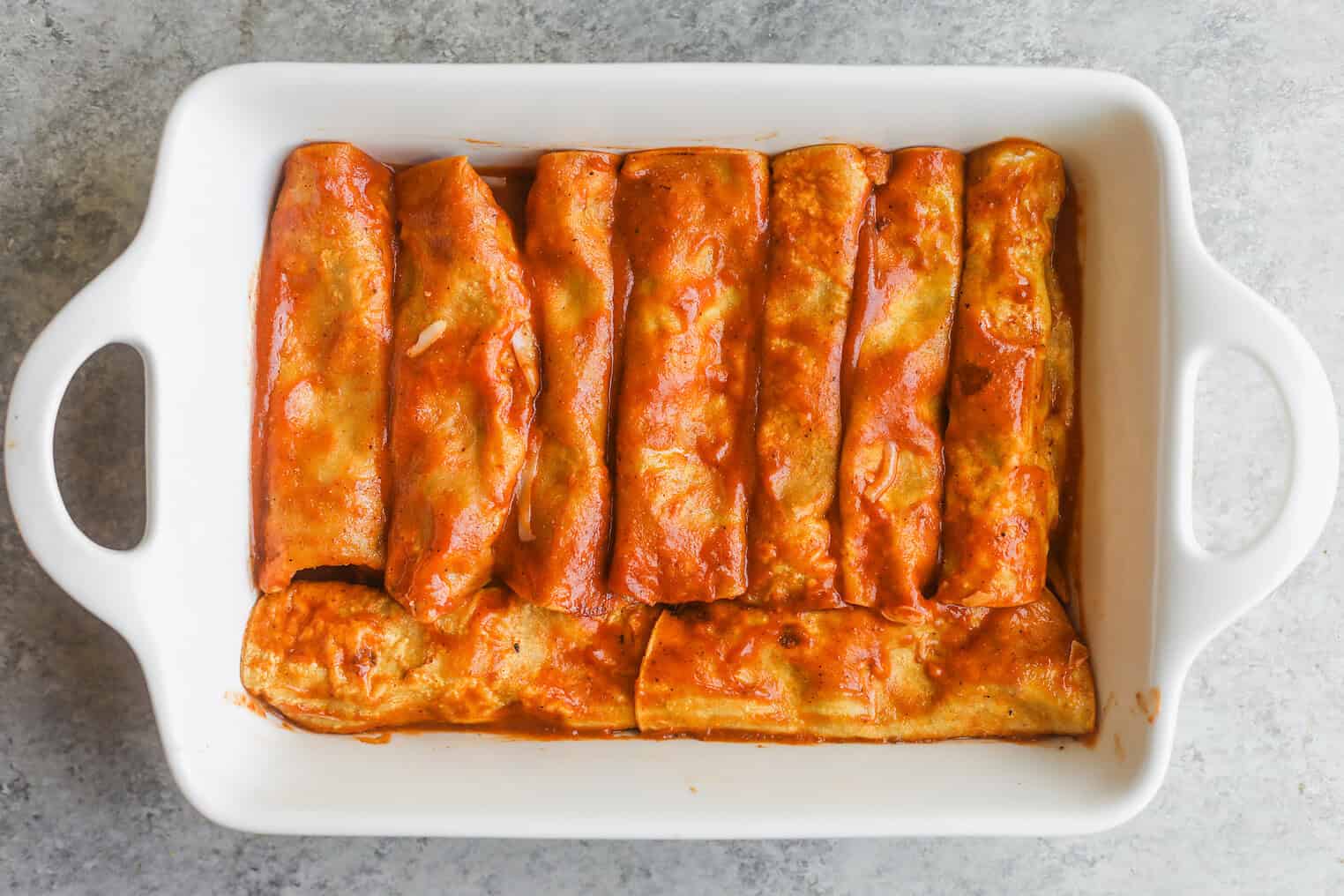 Bean and Cheese Enchiladas 7 A White Rectangular Baking Dish Filled With Eight Bean And Cheese Enchiladas Covered In Red Sauce, Arranged In Rows On A Light Gray Countertop.