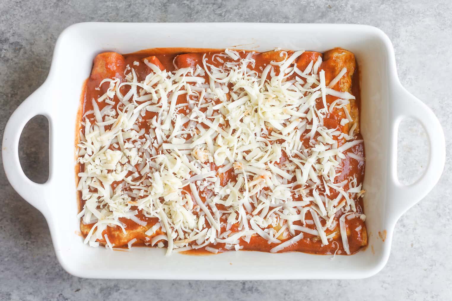 Bean and Cheese Enchiladas 8 A White Baking Dish Filled With Bean And Cheese Enchiladas Topped With Red Sauce And Shredded Cheese, Ready To Be Baked, Sits On A Gray Countertop.