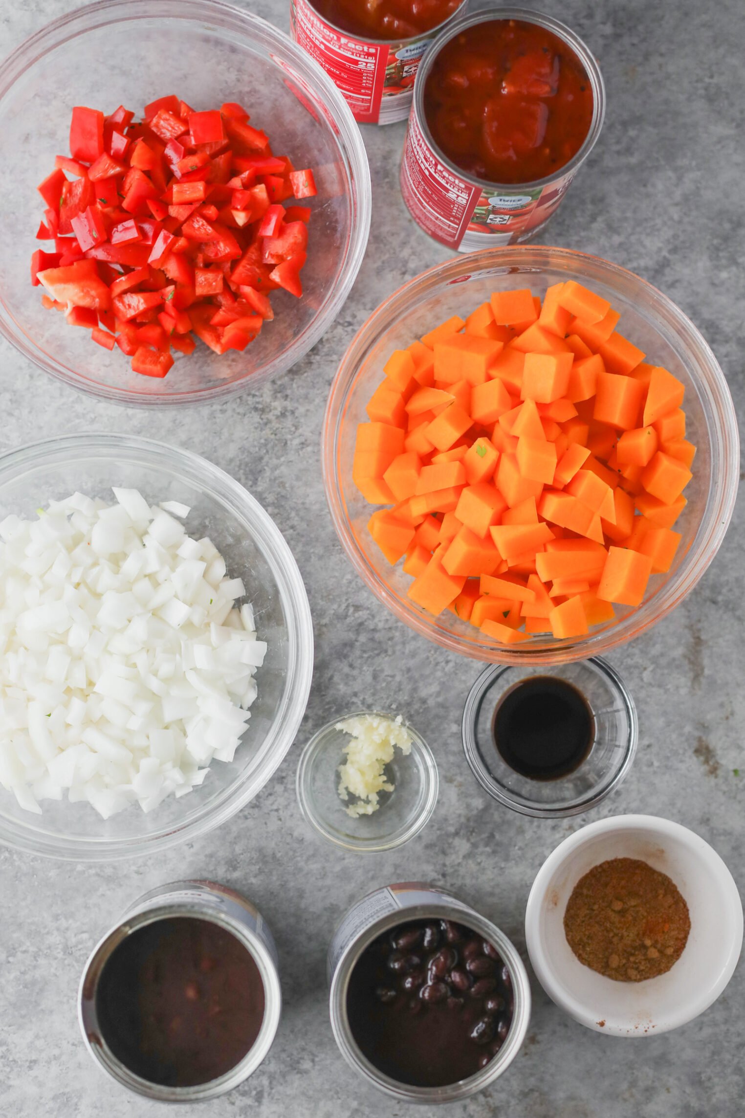 Slow Cooker Black Bean Chili with Butternut Squash 2 Bowls And Cans Filled With Diced Red Bell Pepper, Chopped Carrots, Onion, Minced Garlic, Black Beans, Spices, Sauce, And Canned Tomatoes On A Gray Countertop—Perfect Ingredients For Slow Cooker Vegan Black Bean Chili.