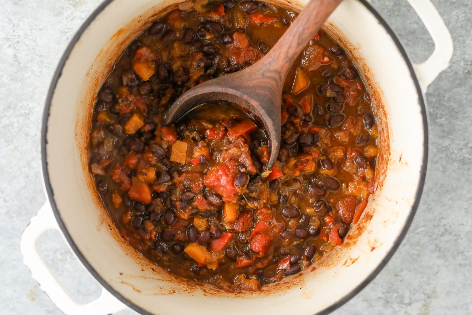 Slow Cooker Black Bean Chili with Butternut Squash 5 A White Pot Filled With Hearty Slow Cooker Black Bean Chili, Featuring Chunky Diced Tomatoes, Carrots, And Onions, Is Being Stirred With A Wooden Spoon. The Stew Appears Well-Cooked And Satisfying.