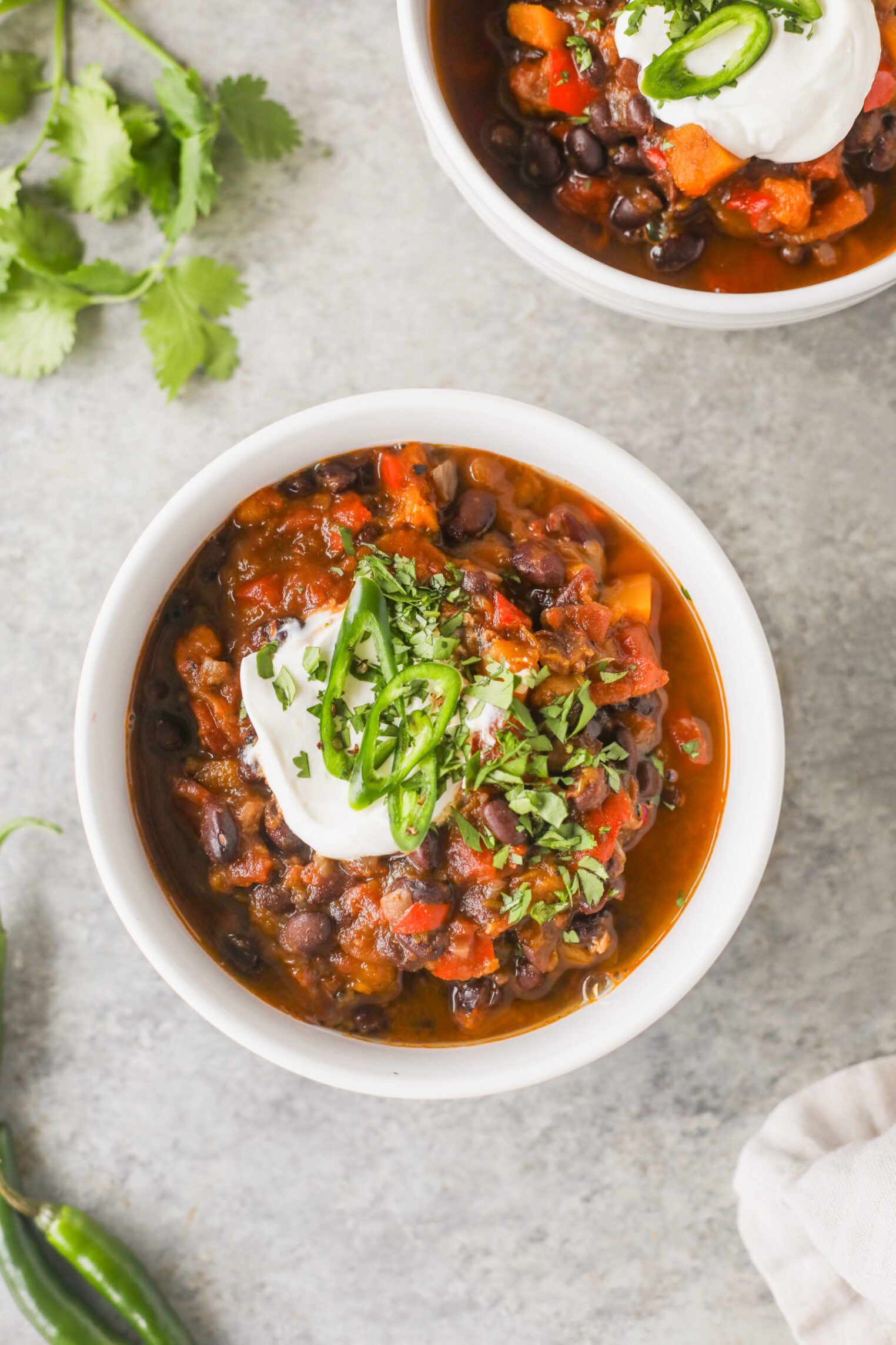 Slow Cooker Black Bean Chili with Butternut Squash 6 A Bowl Of Slow Cooker Black Bean Chili Topped With Sour Cream, Sliced Green Jalapeños, And Chopped Cilantro Sits On A Gray Surface, With Fresh Cilantro And A Green Chili Pepper Nearby. Another Bowl Is Partially Visible.
