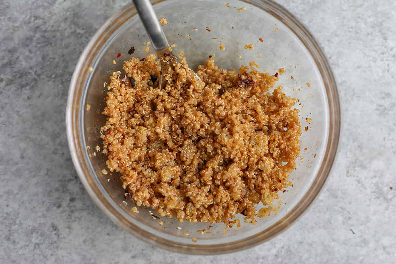 Crispy Quinoa Salad 5 A Glass Bowl Filled With Seasoned, Cooked Quinoa Being Stirred With A Metal Fork, Ready To Become A Delicious Crispy Quinoa Salad On A Light Gray Textured Surface.