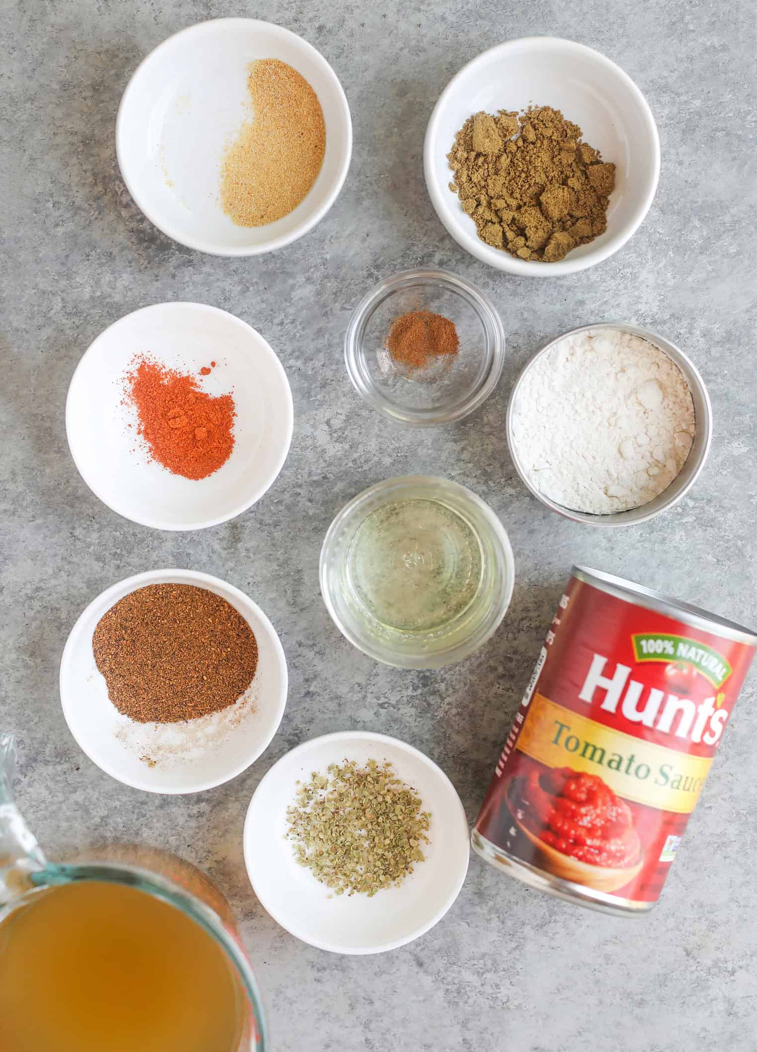 Homemade Enchilada Sauce 3 Various Small White Bowls With Spices, Flour, Oil, And Herbs Sit Next To A Can Of Hunt'S Tomato Sauce And A Measuring Cup Of Broth—Everything You Need For An Easy Homemade Enchilada Sauce, All Arranged On A Gray Countertop.
