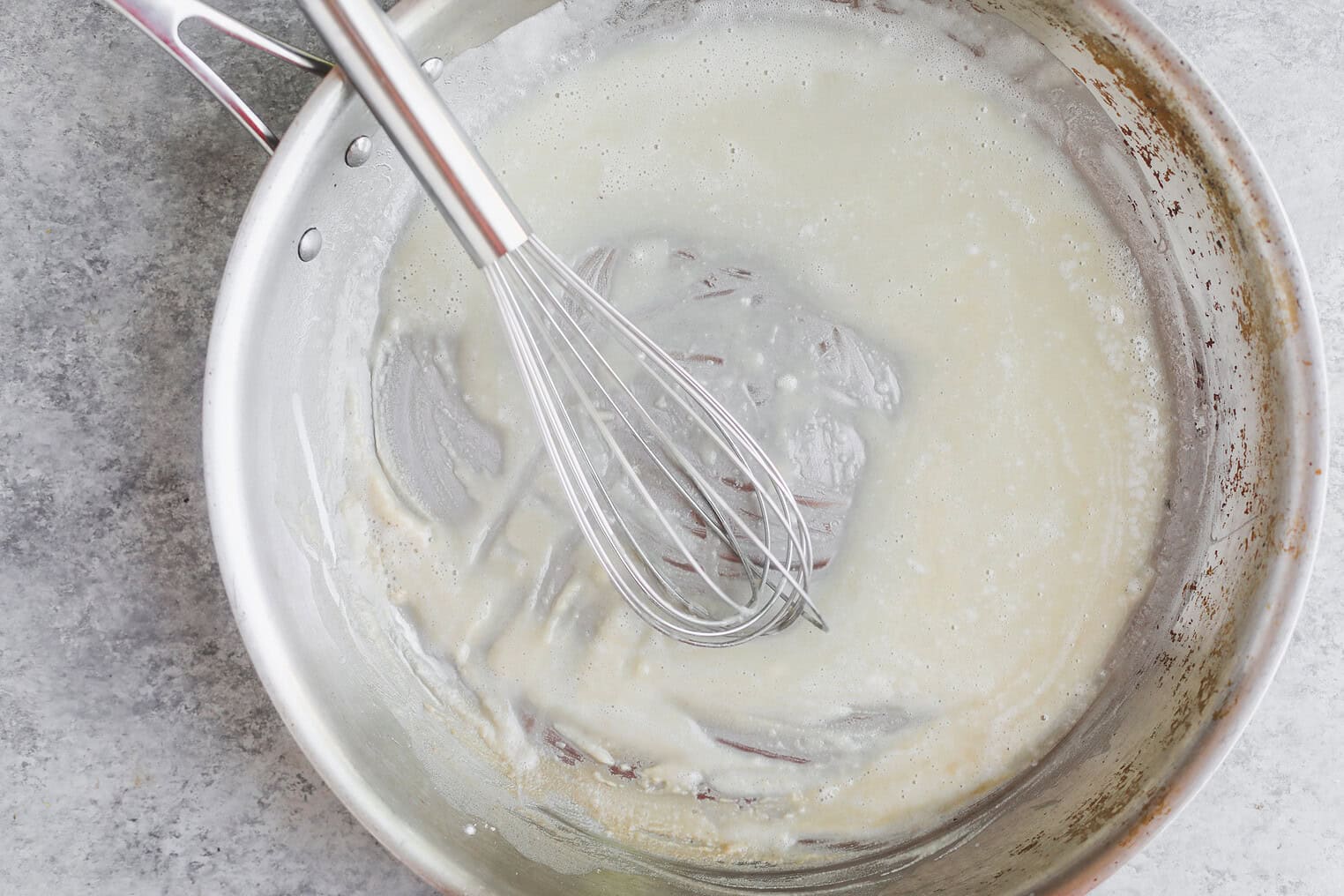 Homemade Enchilada Sauce 5 A Stainless Steel Whisk Stirs A Pale, Thick Mixture In A Metal Saucepan On A Gray Countertop, The Start Of An Easy Homemade Enchilada Sauce Roux.