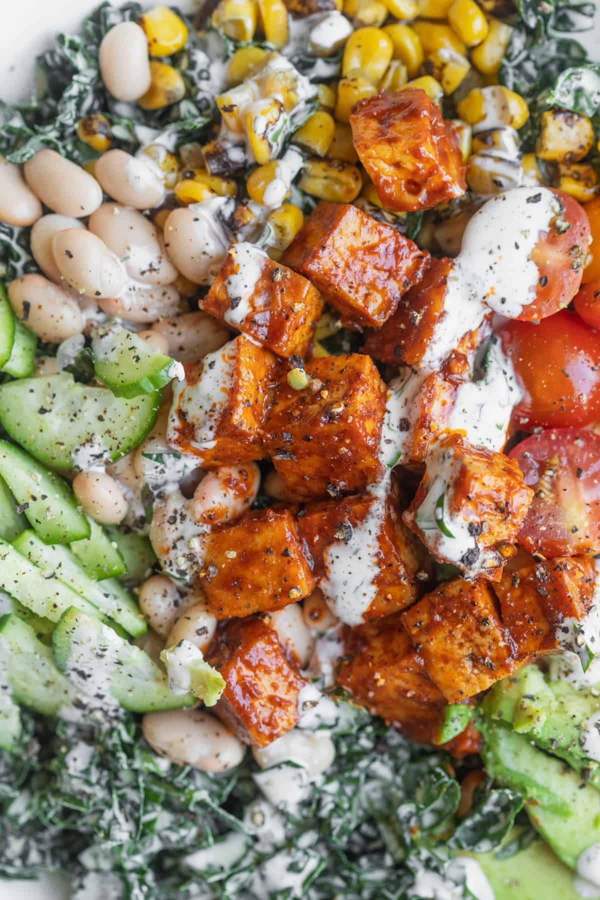 A Close-Up Of A Salad Bowl With Kale, Roasted Corn, White Beans, Cucumber, Tomatoes, Bbq Tofu, And A Drizzle Of Creamy Dressing—An Inspiring Choice For Those Seeking Delicious Vegan Tofu Recipes.