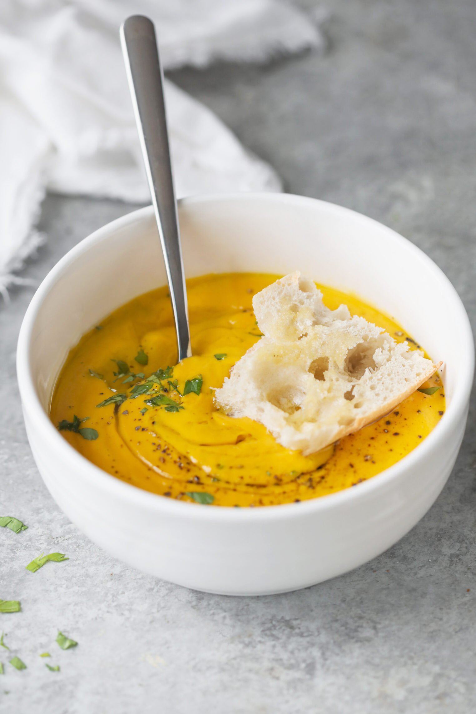 A White Bowl Filled With Creamy Carrot Ginger Soup, Garnished With Herbs And Black Pepper, With A Piece Of Bread Resting On Top And A Spoon Inside. This Immune Boosting Soup Sits On A Gray Surface With A White Cloth Nearby.