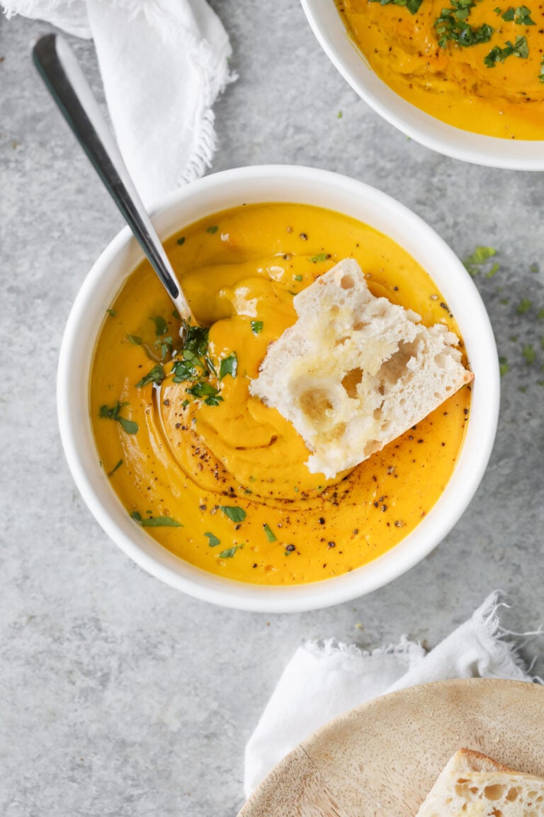A Bowl Of Creamy Orange Carrot Ginger Soup, An Immune Boosting Soup, Is Garnished With Herbs And Black Pepper. A Spoon And Rustic Bread Rest On Top, With A Napkin And Another Plate Nearby On A Gray Surface.