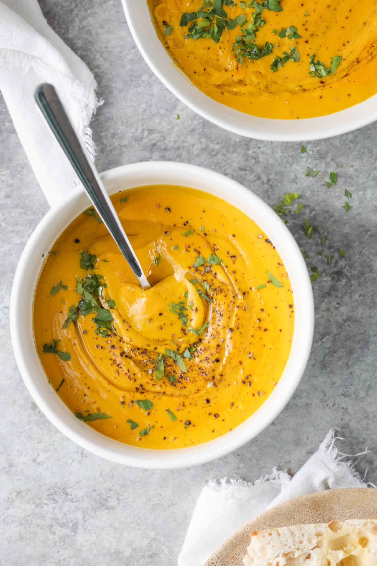 A White Bowl Filled With Creamy Carrot Ginger Soup, Garnished With Chopped Herbs And Cracked Black Pepper, With A Spoon Resting Inside. Another Similar Bowl And A Piece Of Bread Are Partially Visible Beside This Immune Boosting Soup.