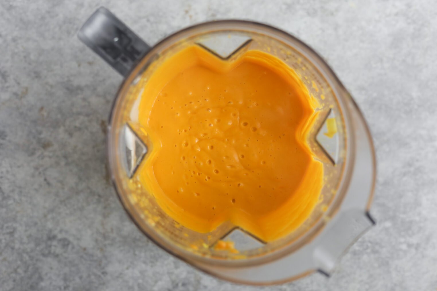 A Top-Down View Of A Blender Containing A Smooth, Creamy Orange Mixture—An Immune Boosting Soup Like Carrot Ginger Soup—With Small Bubbles On The Surface, Set On A Light Gray Countertop.
