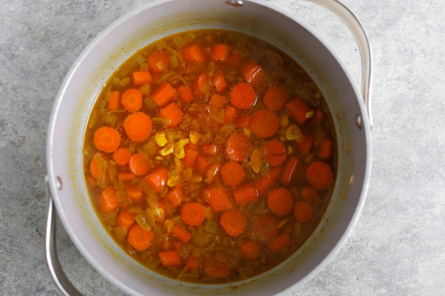 A Pot Filled With Simmering Immune Boosting Soup Containing Sliced Carrots, Onions, And A Golden Broth, Viewed From Above On A Light Gray Surface.