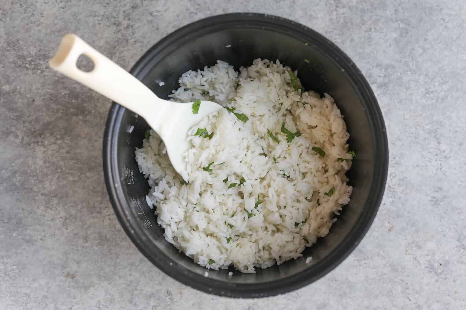 A Black Rice Cooker Pot Filled With Cooked White Rice Mixed With Chopped Herbs, Perfect As A Base For Mango Salsa Bowls, With A White Plastic Rice Paddle Resting Inside. The Pot Sits On A Gray Textured Surface.
