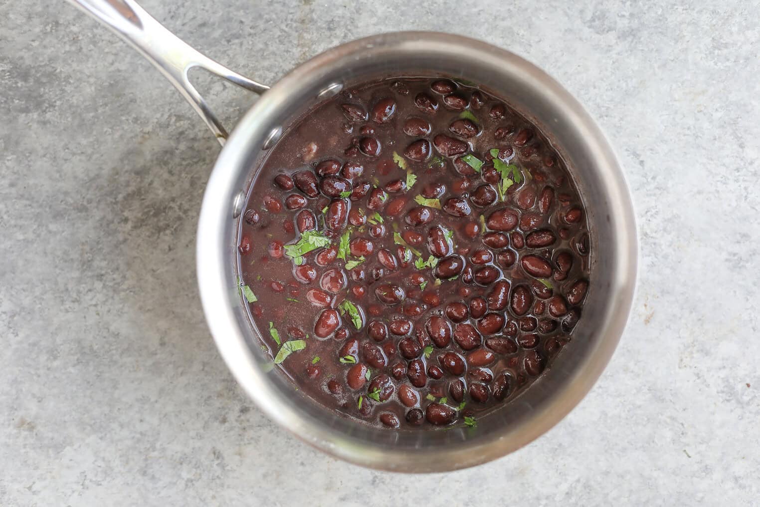 A Stainless Steel Pot Filled With Cooked Black Beans In A Dark Broth, Garnished With Chopped Green Herbs, Sits On A Light Gray Textured Surface—Perfect For Serving Alongside Vibrant Mango Salsa Bowls.