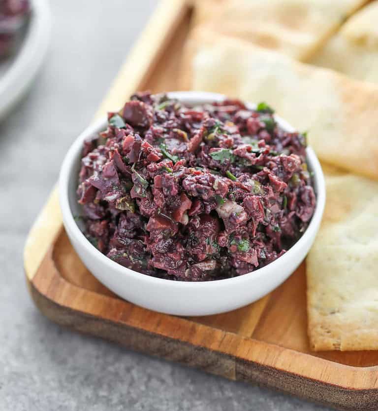 A Small White Bowl Filled With Olive Tapenade, A Healthy Appetizer Option, Sits On A Wooden Board. The Dark Purple Blend Is Mixed With Herbs And Seasonings. Beside The Bowl, Pieces Of Flatbread Await.