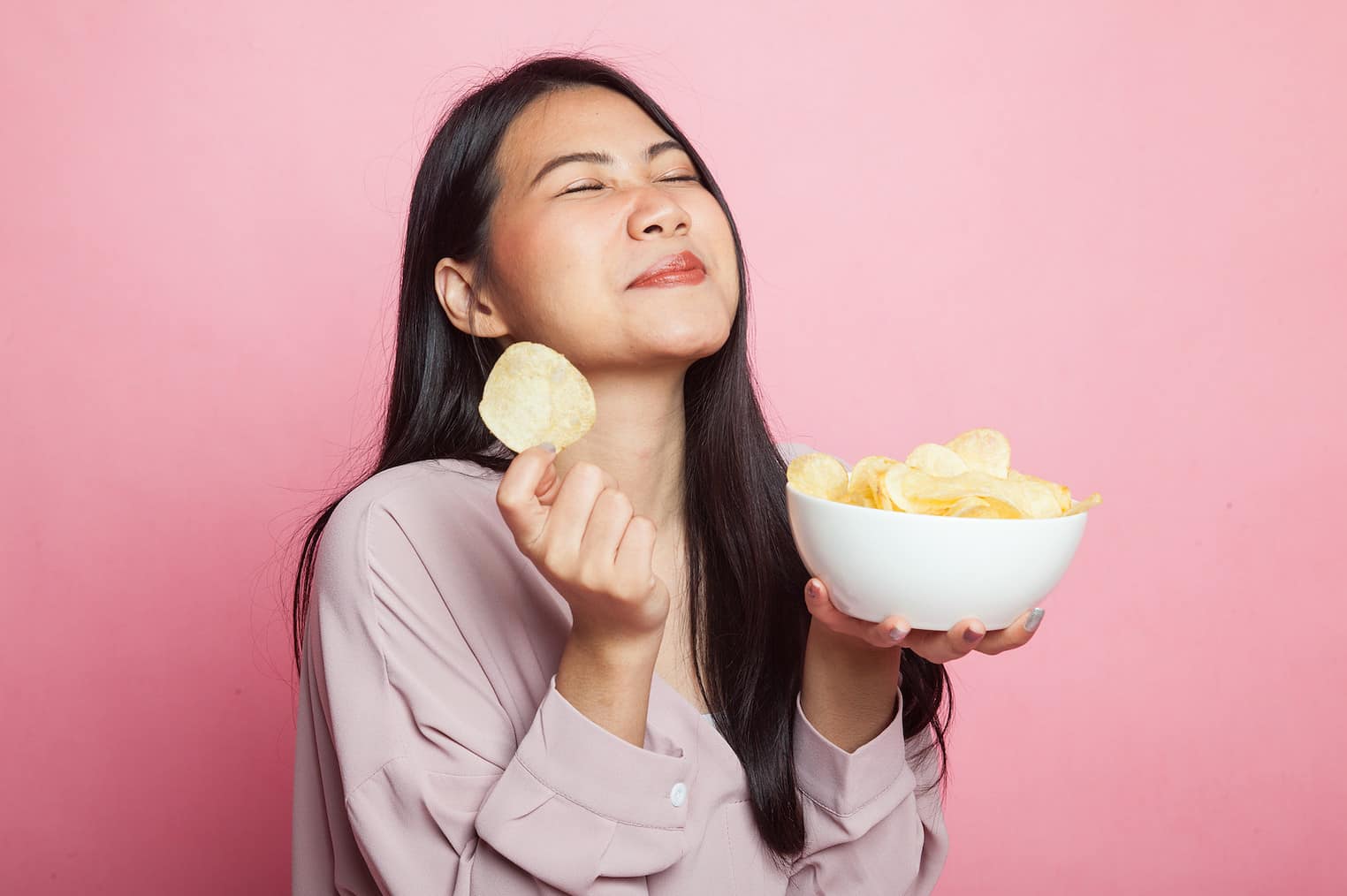 Young Asian woman eat potato chips on pink background