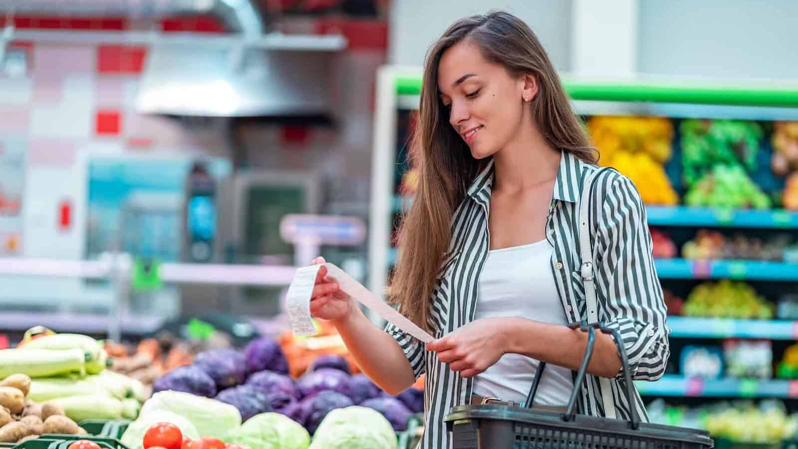 Young woman with shopping basket checks and examines a sales receipt after purchasing food in a grocery store. Customer buying products at supermarket