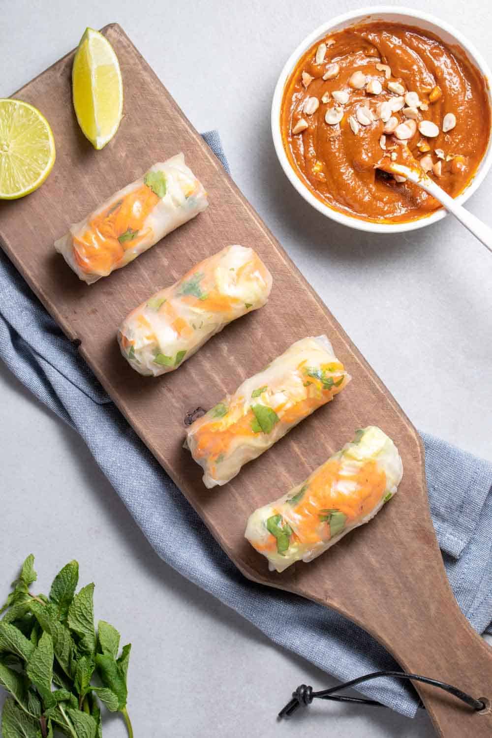 Top View Of Spring Rolls On Serving Board With Lime Wedge And Spicy Peanut Sauce In A Bowl 